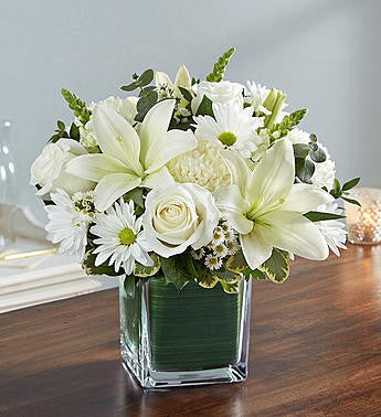 A square clear glass cube with a leaf wrapped in the bottom and filled with beautiful assorted white blooms. White lilies, snapdragons, daisies, roses and accents of greenery.  Vase and/or flower variety will vary depending on availability. Arrangement will be as close as possible, but may not be exact.