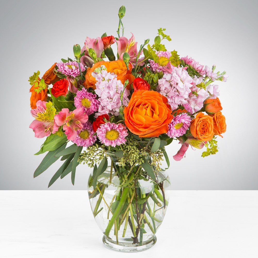 Flowers in the colors of orange and pastel pink with roses, stock, asters, alstromeria and carnations.   Vase and/or flower variety will vary depending on availability. Arrangement will be as close as possible, but may not be exact.