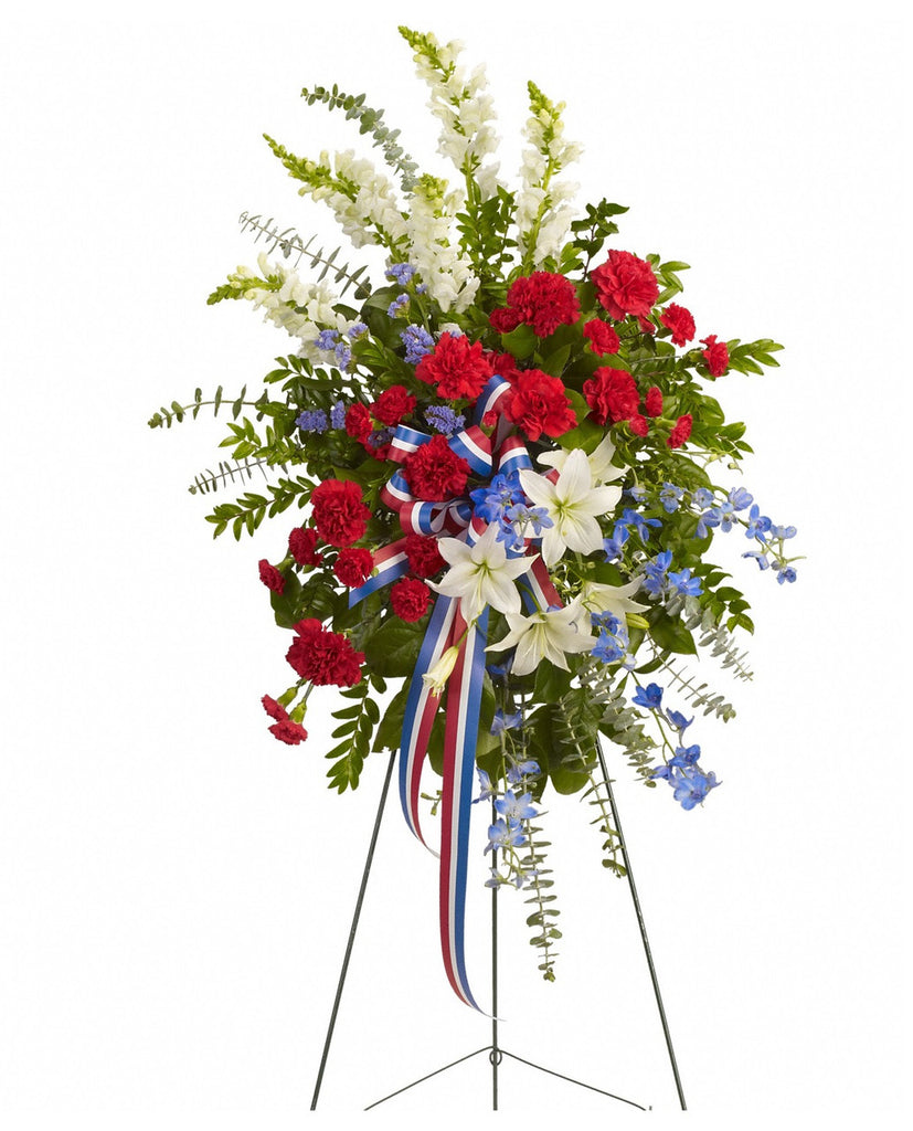 Floral arrangement with red, white, and blue flowers and ribbons on a stand against a white background