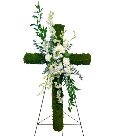 A floral arrangement featuring a moss-wrapped cross with a white flower spray and green foliage in the center.