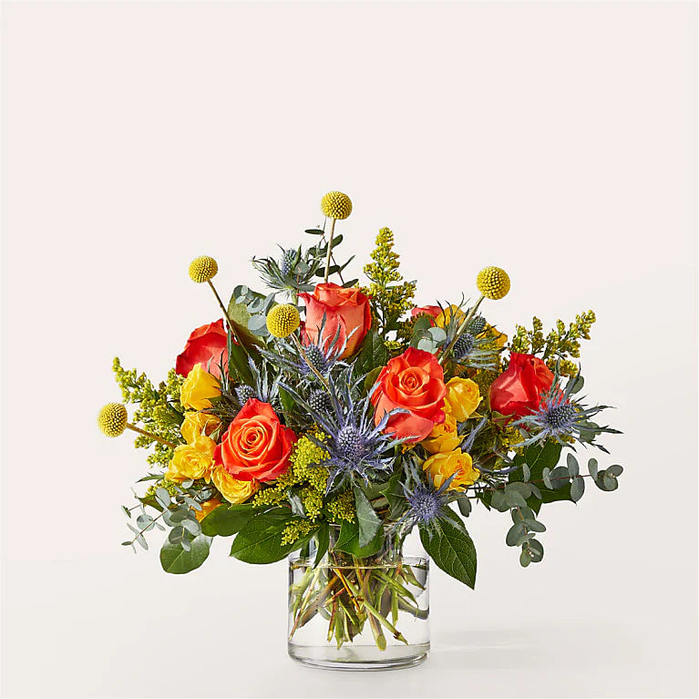 Bouquet of orange roses, yellow flowers, blue thistle and greenery in a clear vase on a light background