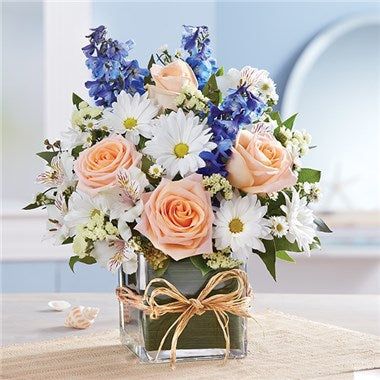 A clear square glass vase wrapped with a green leaf and filled with white daisies, blue delphinium and peach roses.  Vase and/or flower variety will vary depending on availability. Arrangement will be as close as possible, but may not be exact.