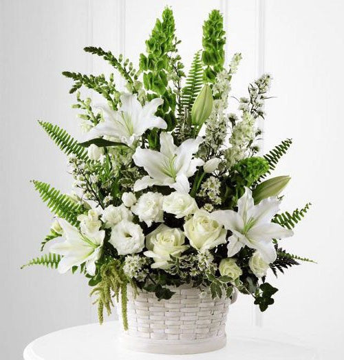 An all white floral arrangement in a white wicker basket. Includes bells of ireland, white snapdragons and lush with the lilies, roses, lisianthus and filler.   Basket and/or flower variety will vary depending on availability. Arrangement will be as close as possible, but may not be exact.