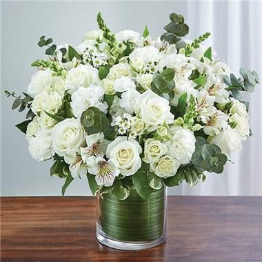 An arrangement of white roses, snapdragons, alstroemeria, carnations, eucalyptus in a clear glass cylinder vase with a green leaf ribbon.   Vase and/or flower variety will vary depending on availability. Arrangement will be as close as possible, but may not be exact.