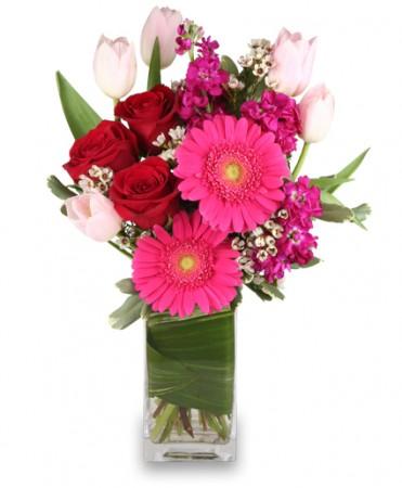 A rectangular clear vase wrapped with a green leaf and filled with tulips, roses, stock and gerbera daisies. Pinks and reds.    Vase and/or flower variety will vary depending on availability. Arrangement will be as close as possible, but may not be exact. 