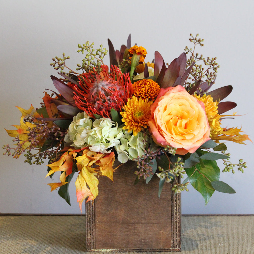 A square wooden box filled with fall colors of pin cushion protea, marigolds, roses, hydrangeas, oak leaves and eucalyptus.   Box and/or flower variety will vary depending on availability. Arrangement will be as close as possible, but may not be exact.